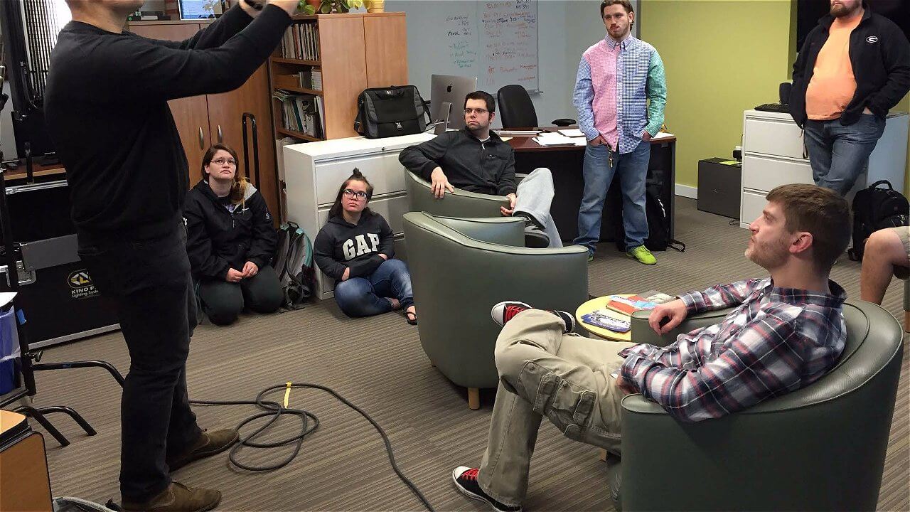 A group of people in a casual office setting, some sitting on chairs while others are standing, engaged in discussion. One person is holding a camera or device, directing attention towards the group. The atmosphere appears collaborative and interactive.