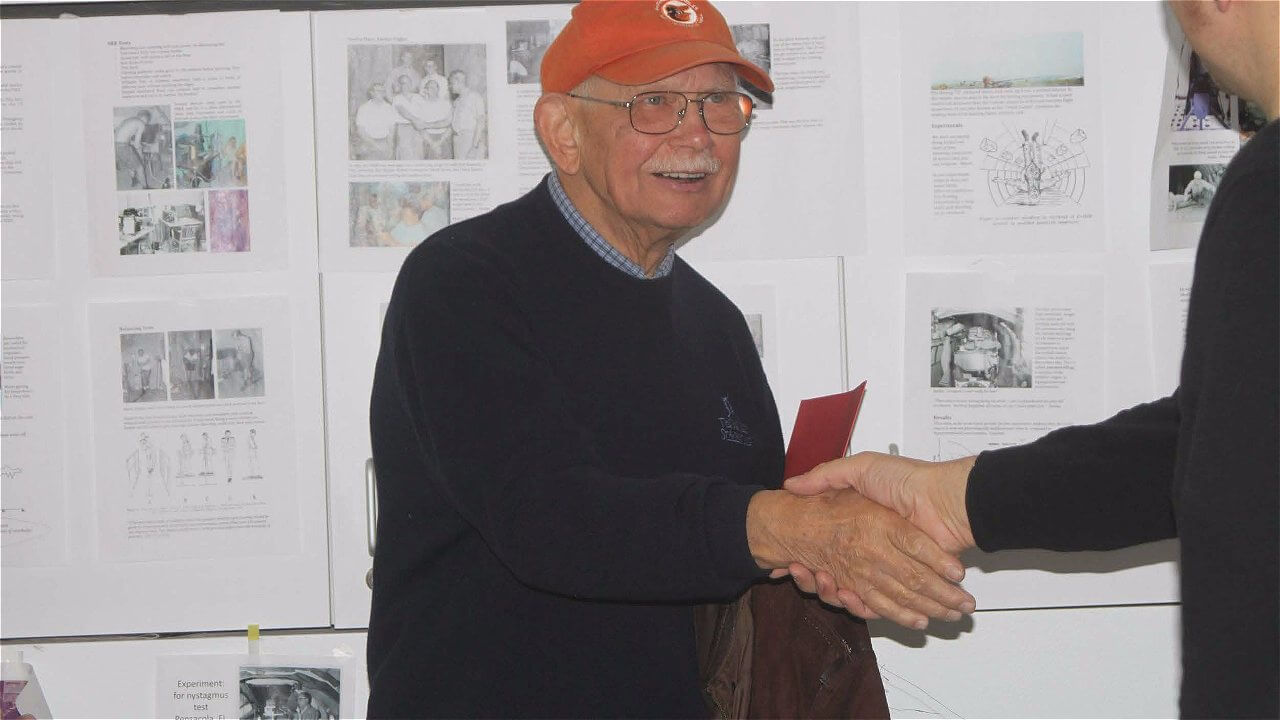An elderly man with glasses and a bright orange cap is smiling and shaking hands with another person. Behind them, there are several sheets of paper displayed on a wall, featuring various images and text.