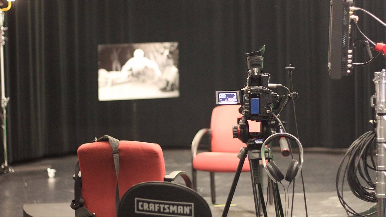 A camera set up on a tripod facing two red chairs in a studio setting, with a black backdrop and a framed image hanging on the wall behind them. There are studio lights and audio equipment visible in the scene.