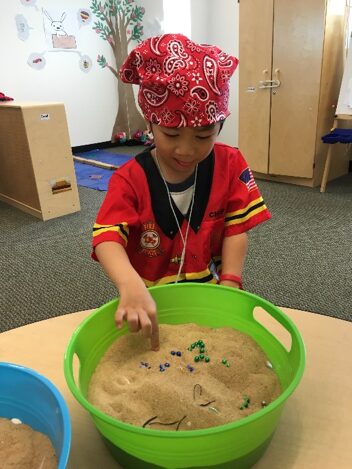 A young child wearing a red bandana and a colorful shirt is playing with a green bowl filled with sand. The child is reaching into the sand, which contains various small objects, possibly for exploration or a sensory activity. The background features a classroom setting with a wooden structure and colorful decorations.