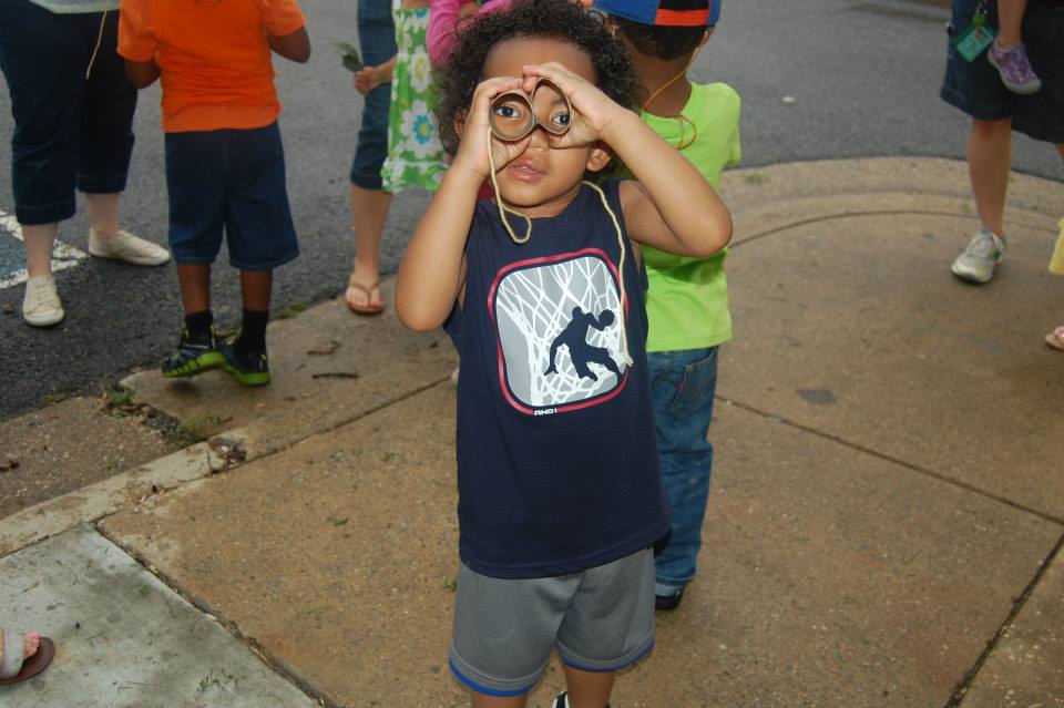 A young child wearing a dark t-shirt and shorts is holding a pair of glasses in front of their face, pretending to look through them. In the background, other children and adults can be seen, some wearing colorful clothing. The setting appears to be outdoors on a paved surface.