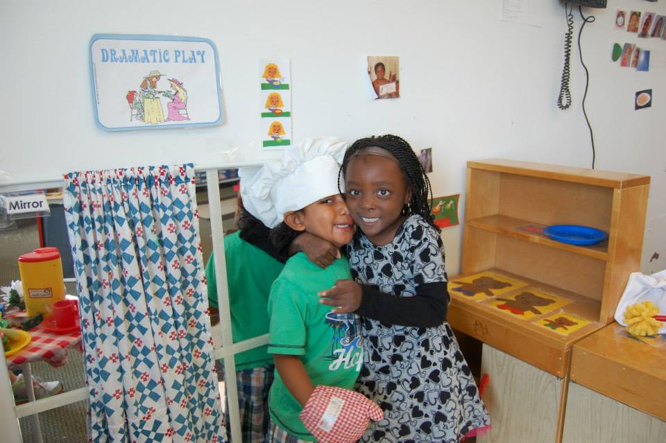 Two children are smiling and embracing each other in a playful classroom setting. One child is wearing a chef's hat and a green shirt, while the other wears a black and white patterned dress. In the background, there are colorful play items and a sign that reads 