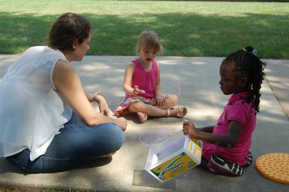A woman and two young girls are sitting on a concrete surface outdoors. The woman, wearing a white top and blue jeans, is engaged in conversation with the girls, who are dressed in pink. One girl is sitting cross-legged, while the other is kneeling and holding a box of crayons. The background features a grassy area.