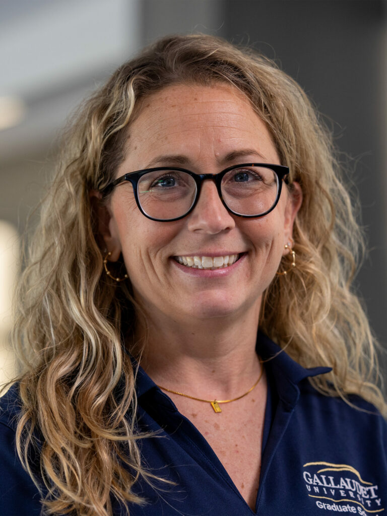 A smiling woman with long, curly blonde hair wearing glasses and a navy blue shirt with a logo.