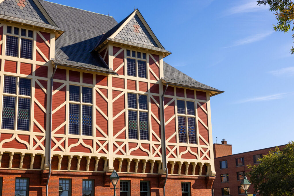 A close-up view of a historic brick building featuring intricate wooden framing, large windows, and a steep, gabled roof. The building is set against a clear blue sky, with a glimpse of another structure in the background and greenery in the foreground.
