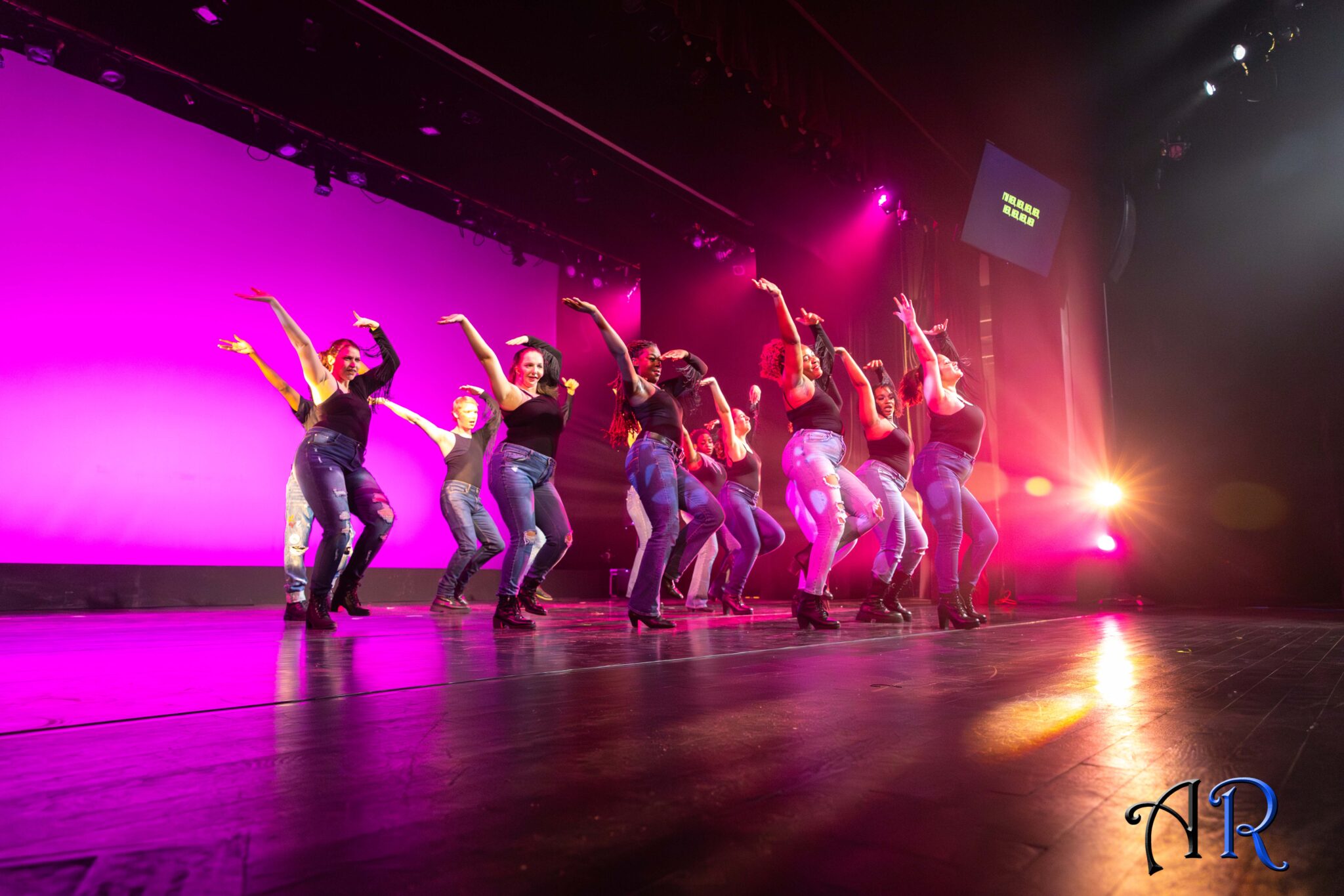 Pink background, 13 dancers in jeans and black one sided sleeve leotard with tassels hanging from under sleeve. Dancing arms up