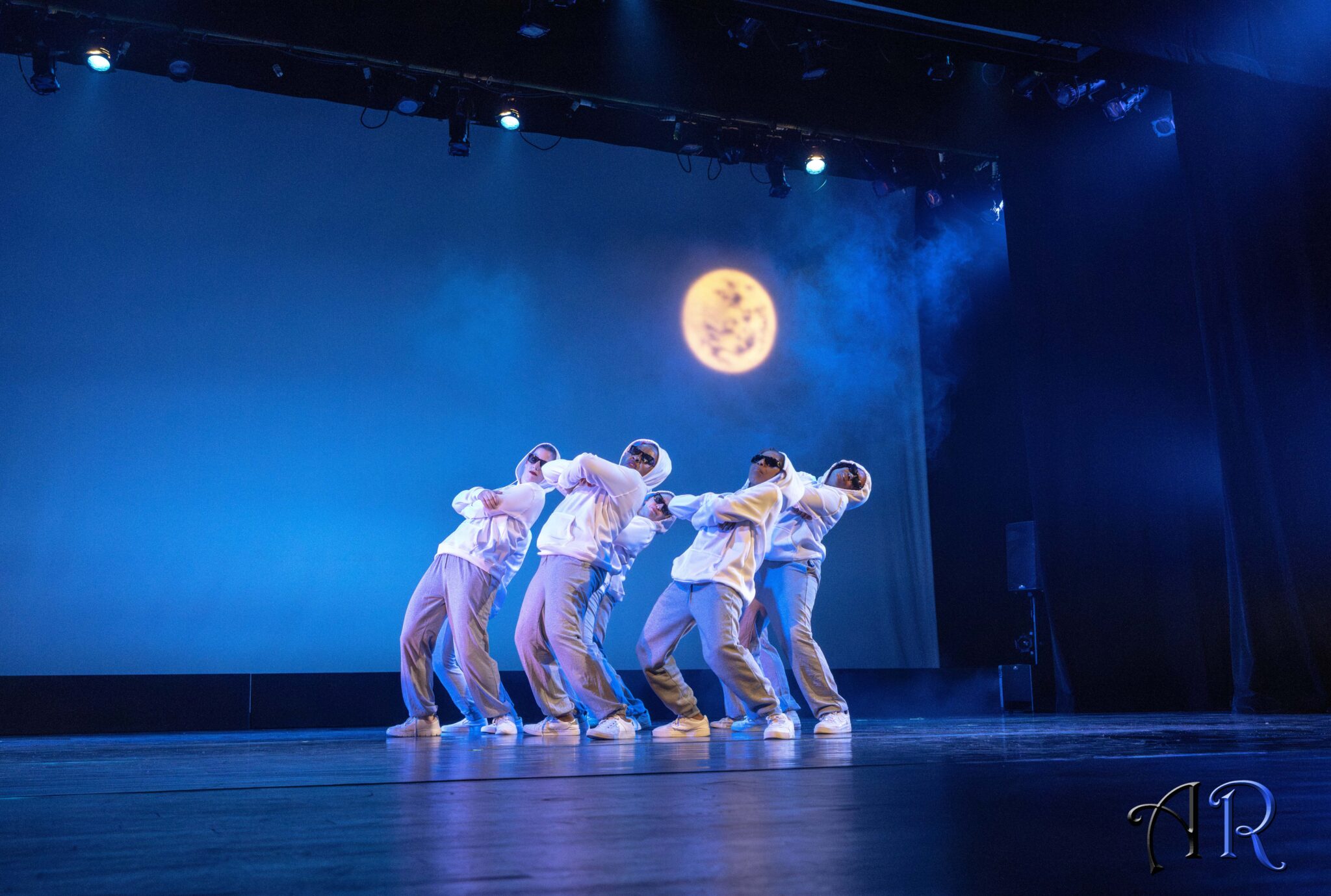 Six dancers with blue night background and shiny white moon, all dancers in sun shades with white hoodie and grey sweatpants, leaning back.