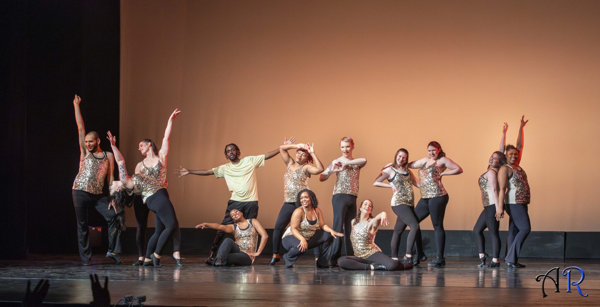 Gold background , 13 dancers wearing gold tops and black pants posing at the end of performance, most standing up, 3 sitting on the floor in the front