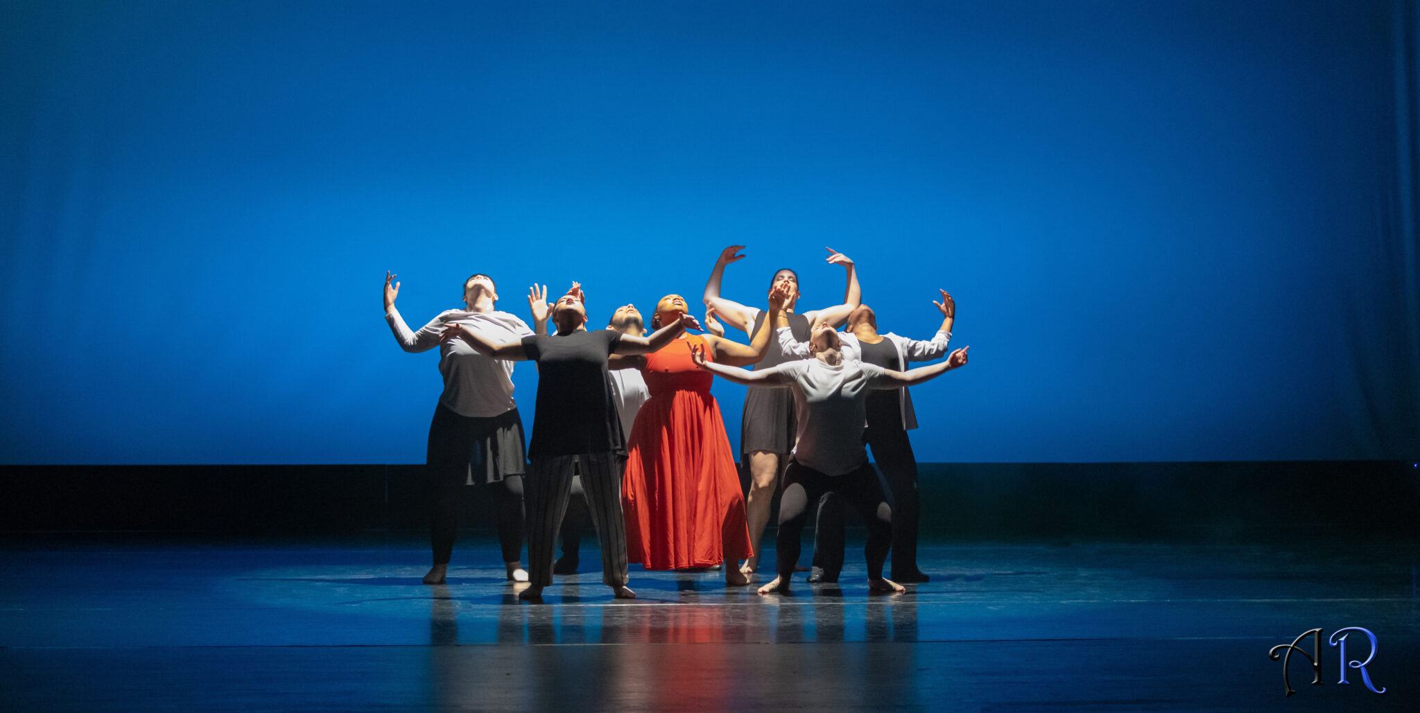 Blue background, 6 dancers wearing black and white clothes circling a woman with a red dress, all pleaing, hands up and looking up to light on them