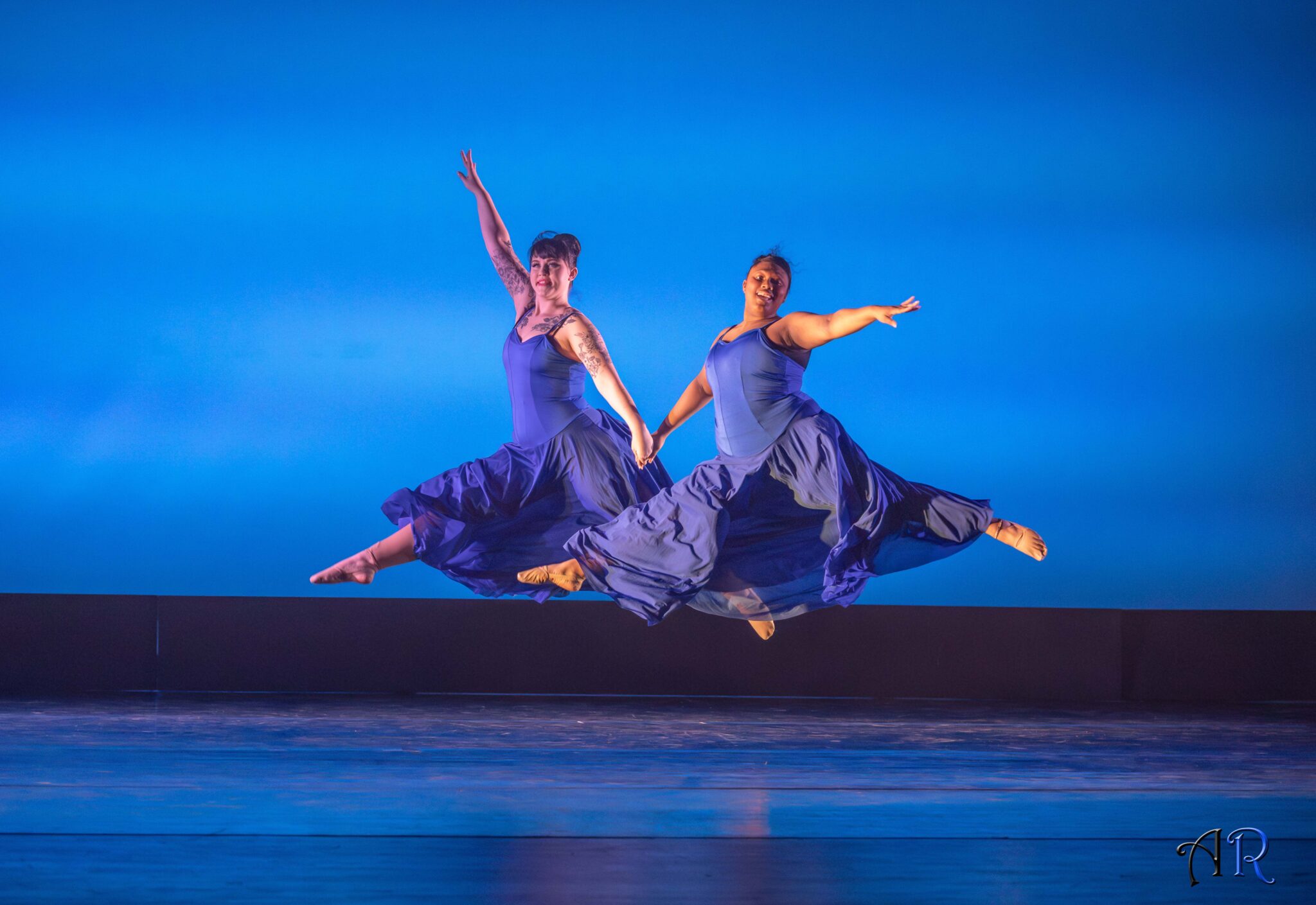 Blue background, two dancers leaping in air holding hands , blue dresses