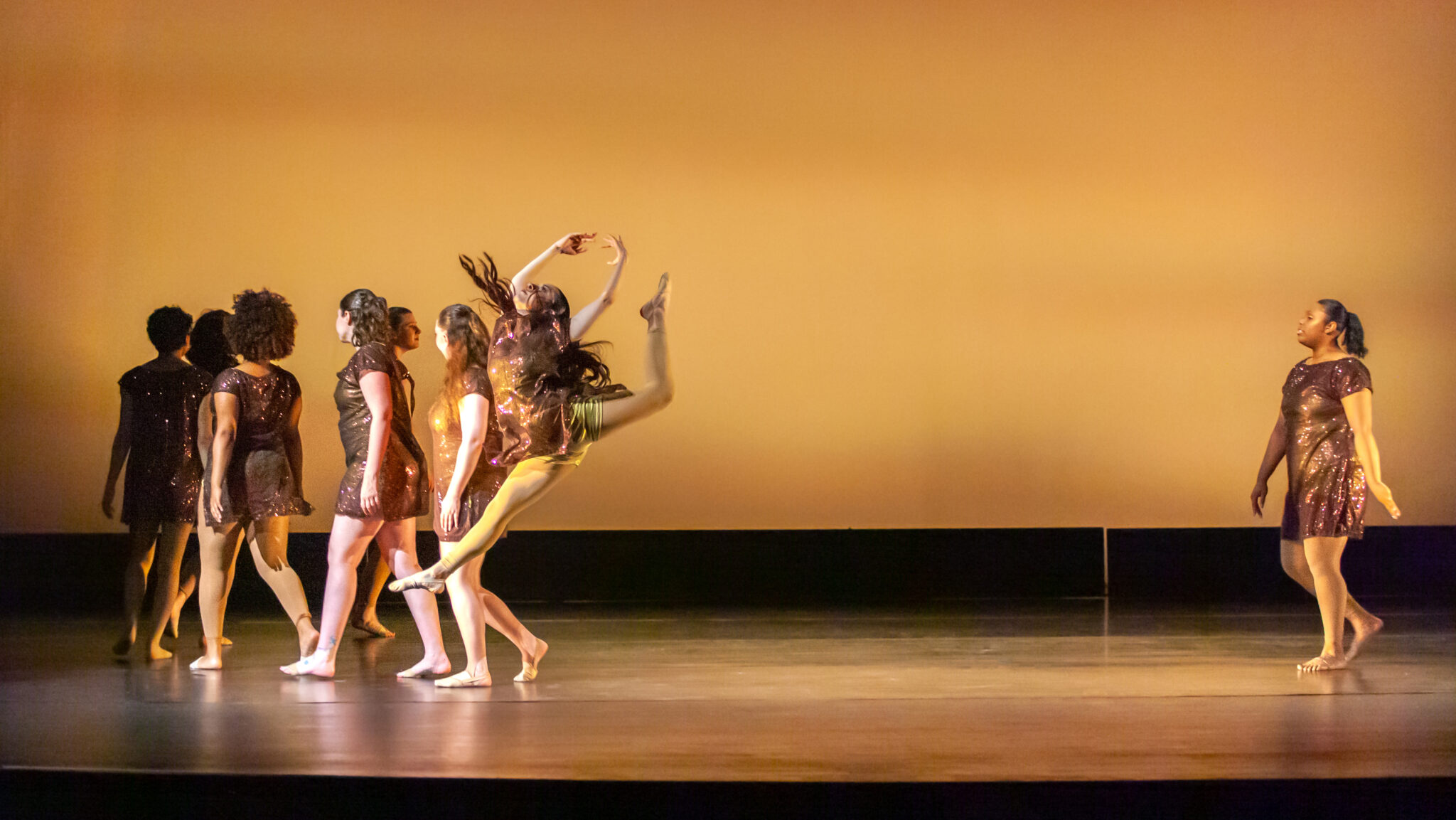 Gold background, dancers in gold dress one leaping in the front, other girls in a group in the back