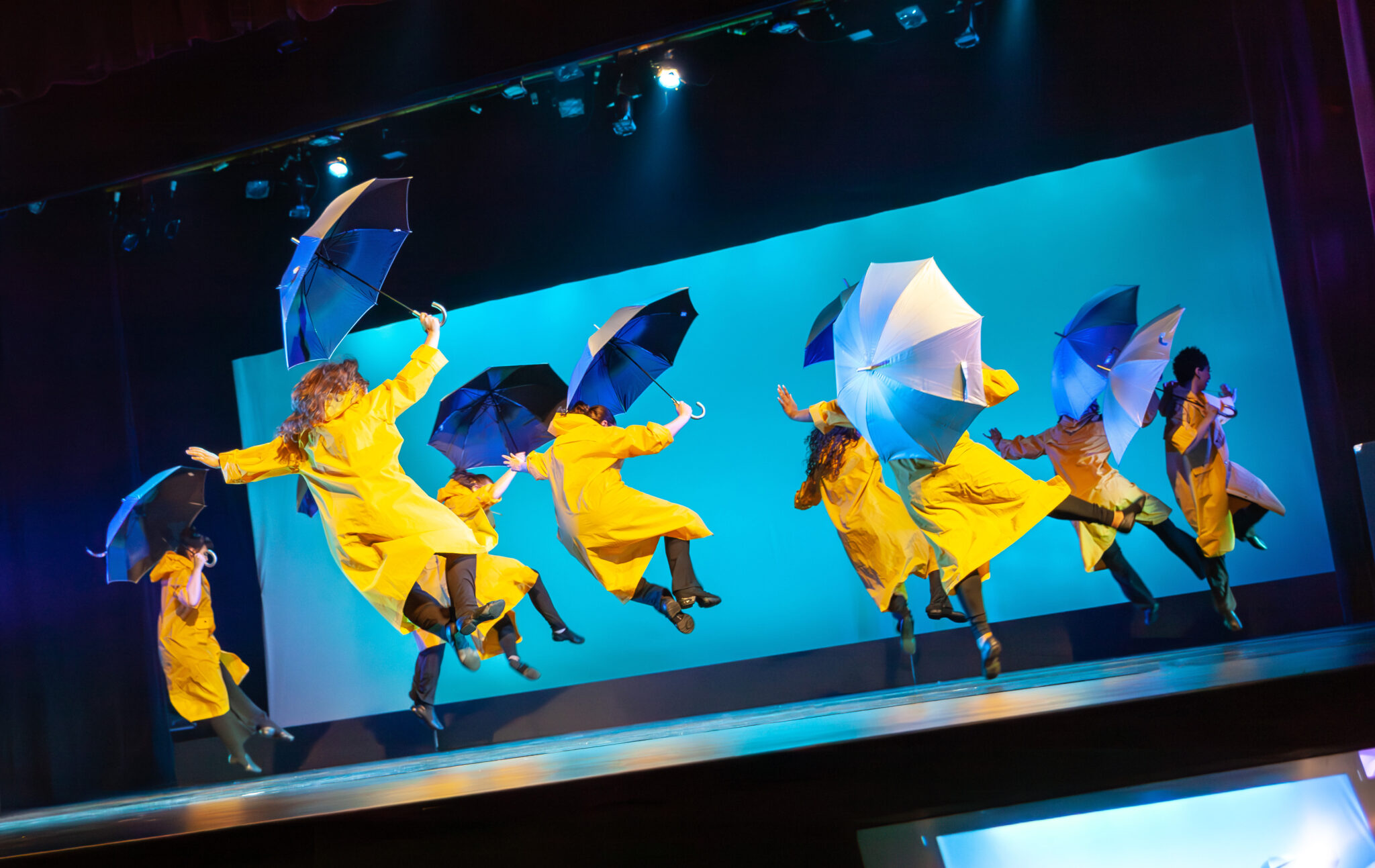 Dancers in yellow rain coat with umbrellas, blue background jumping on air
