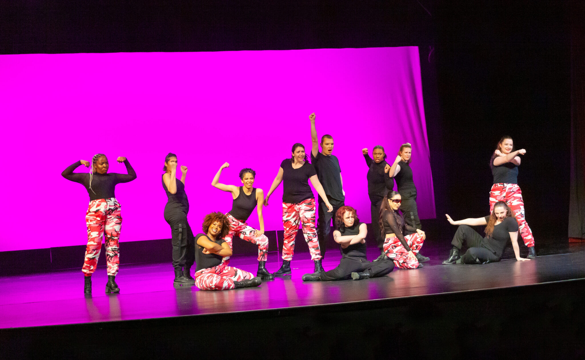 Purple background, dancers posing with black shirt and pink camouflage pants, some have black pants and sitting non the ground in front