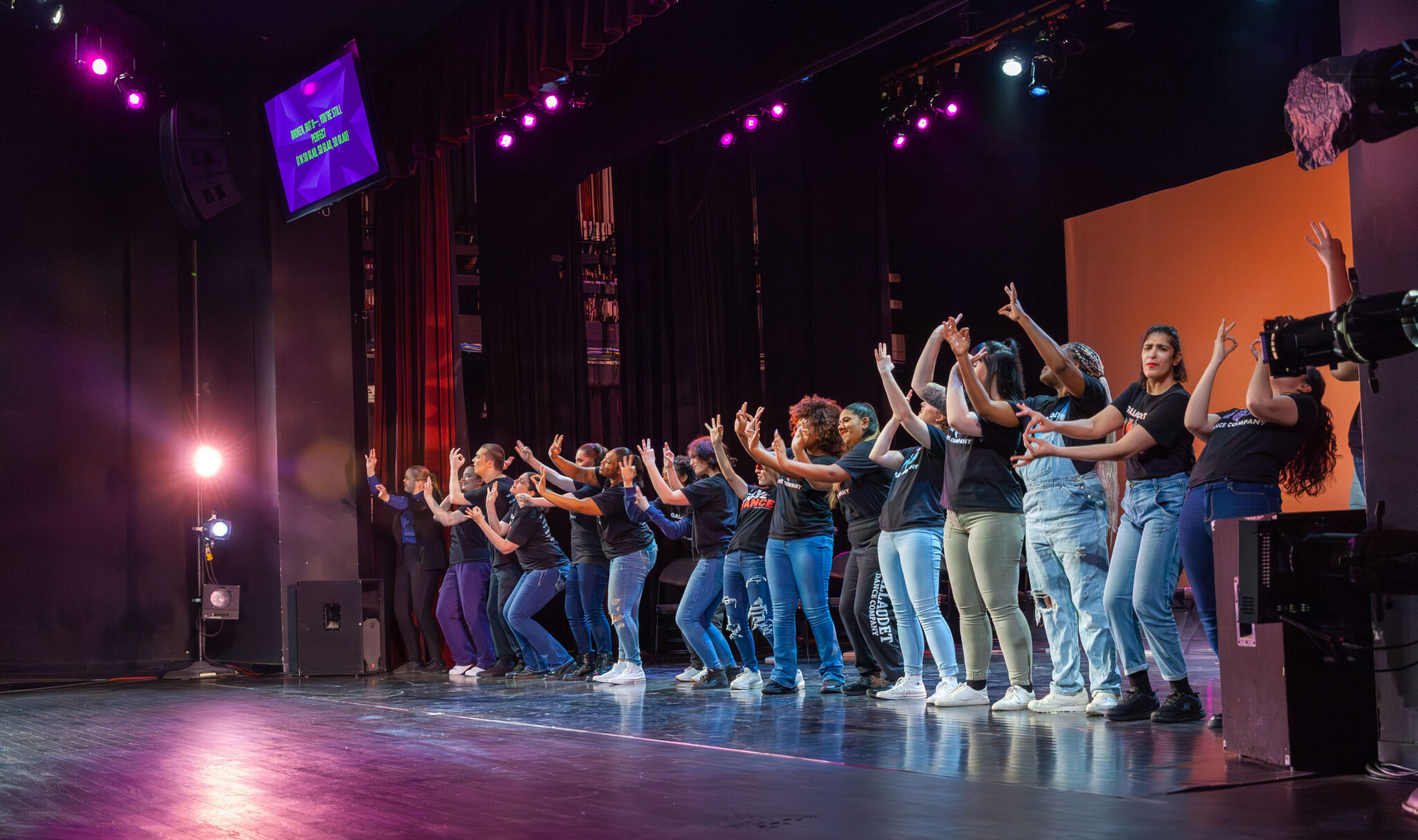 All dancers in a line in the front with blue jeans and black gdc shirt, all signing “perfect” “9-9”
