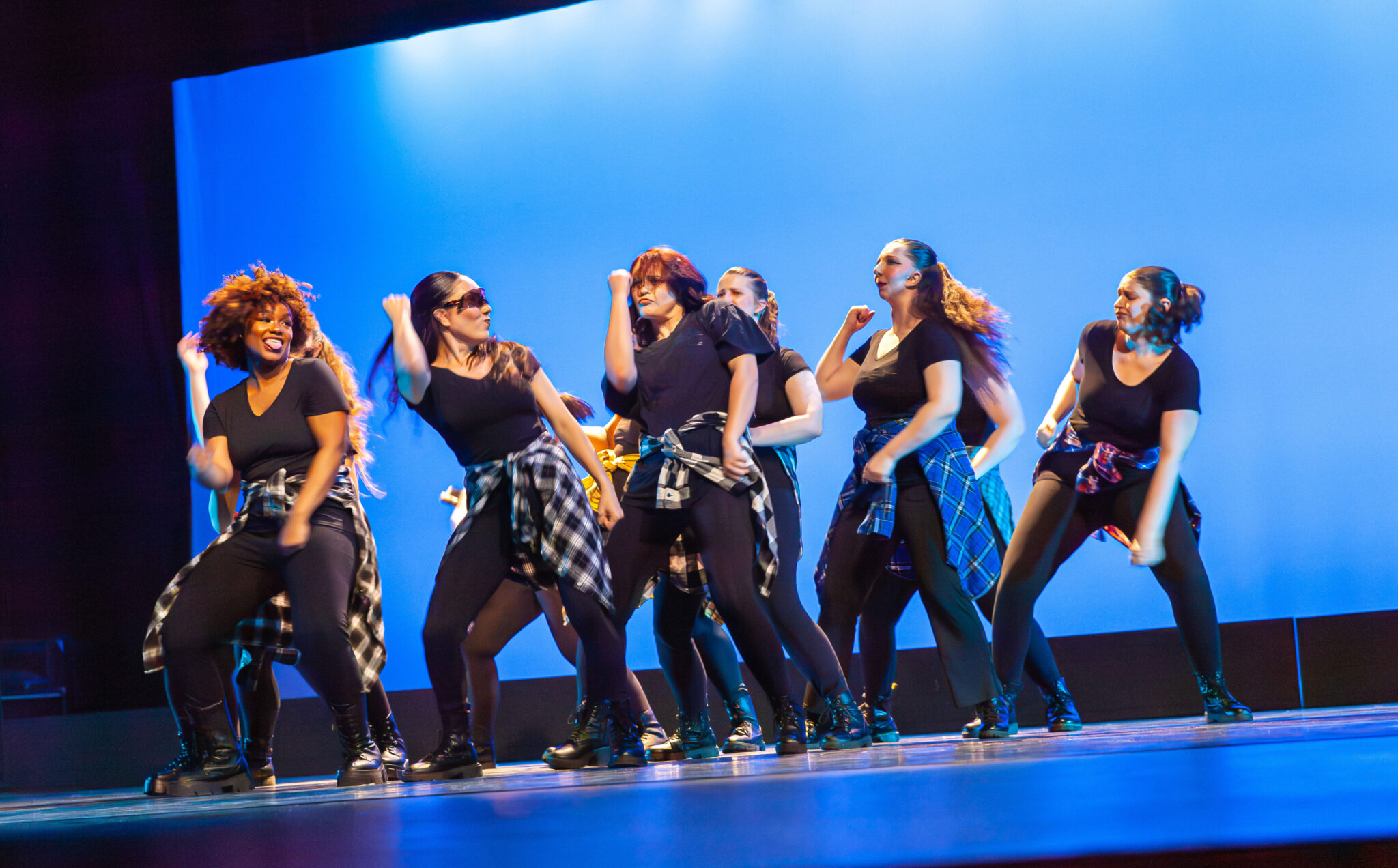 Blue background, dancers in a triangle formation, dressed in all black with flannel tied on waist.
