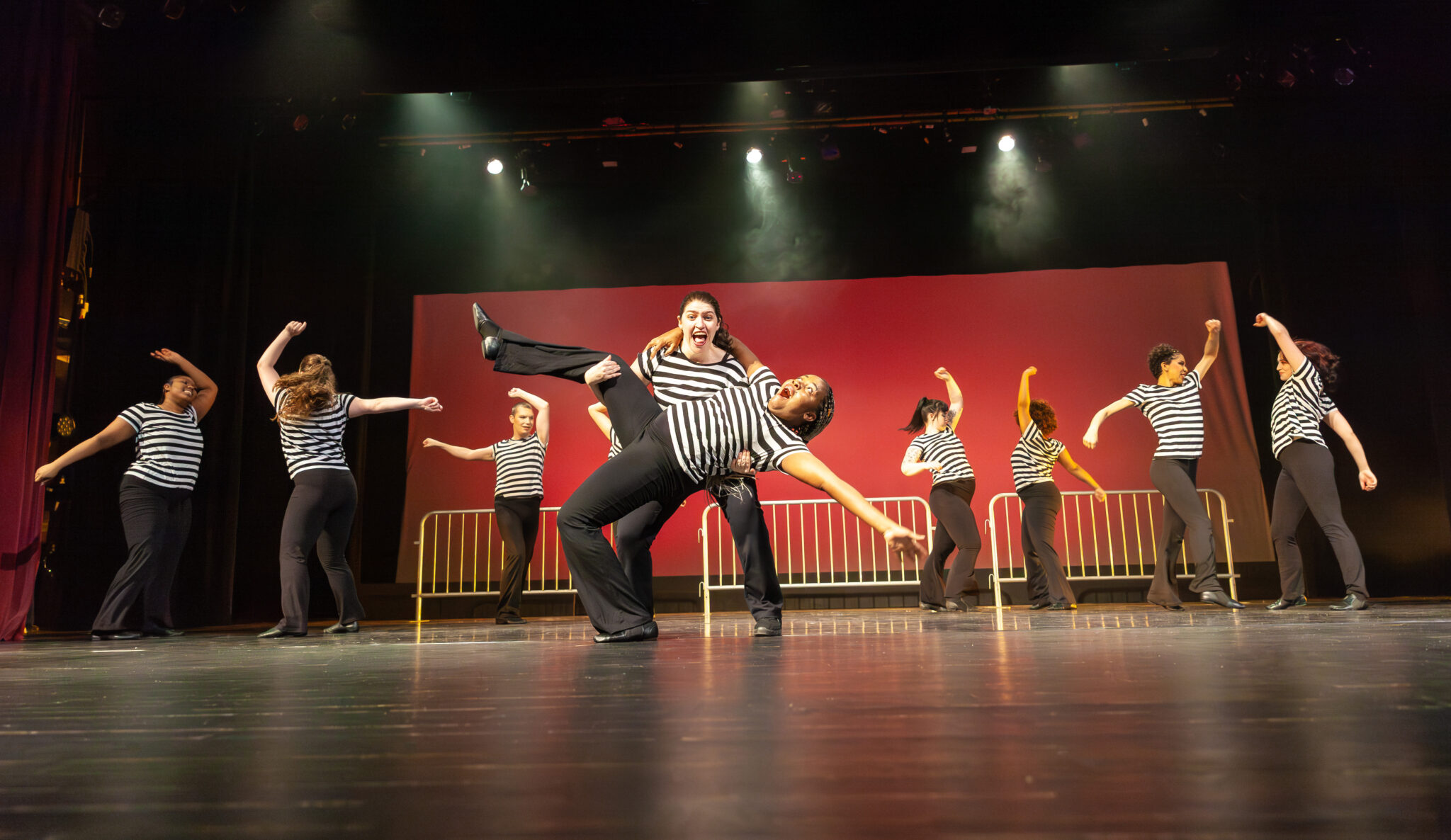 Red background, dancers wearing striped black/white top with black pants and prison like setting