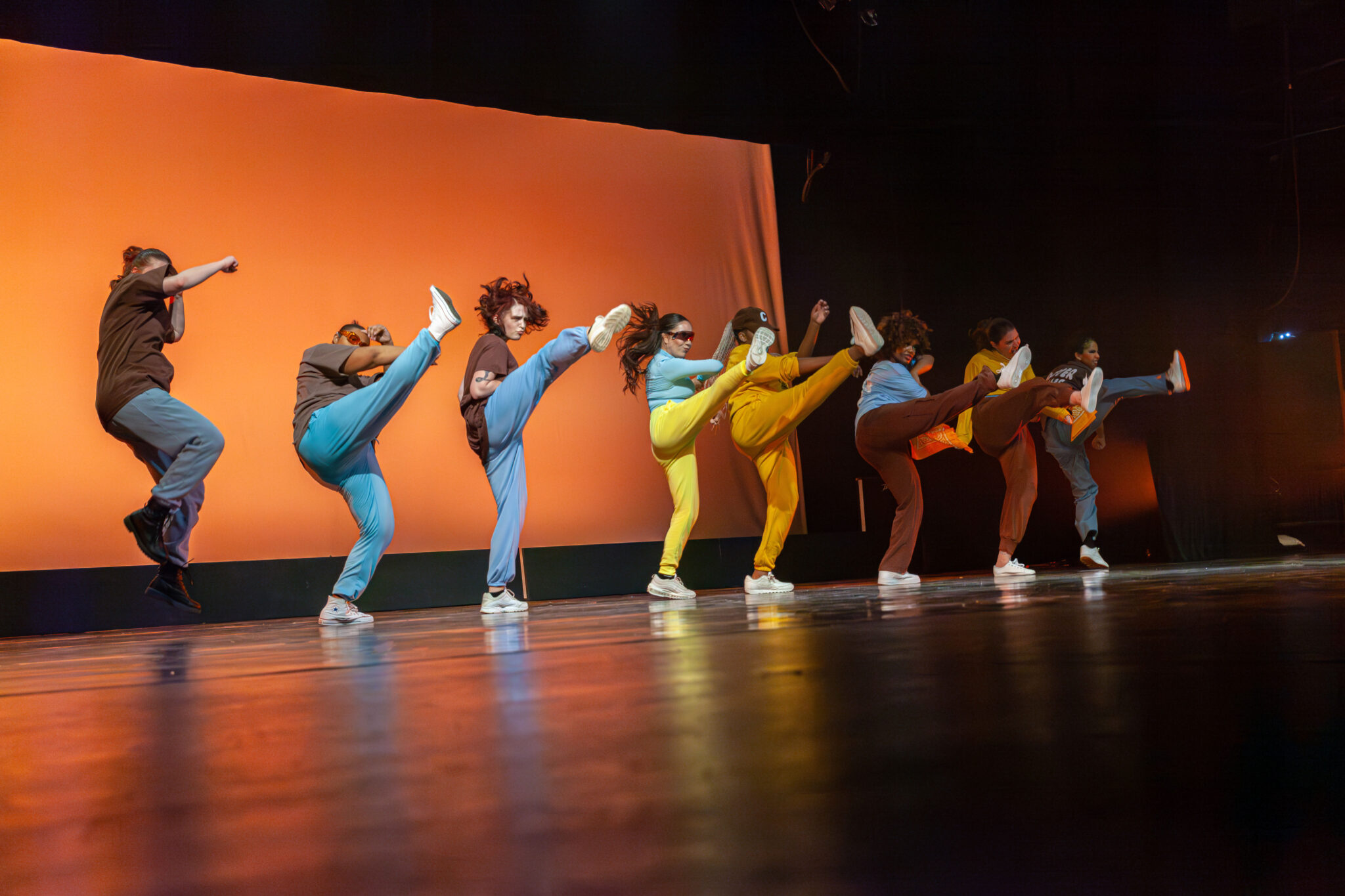 Yellow background, line of dancers all kicking their right leg out