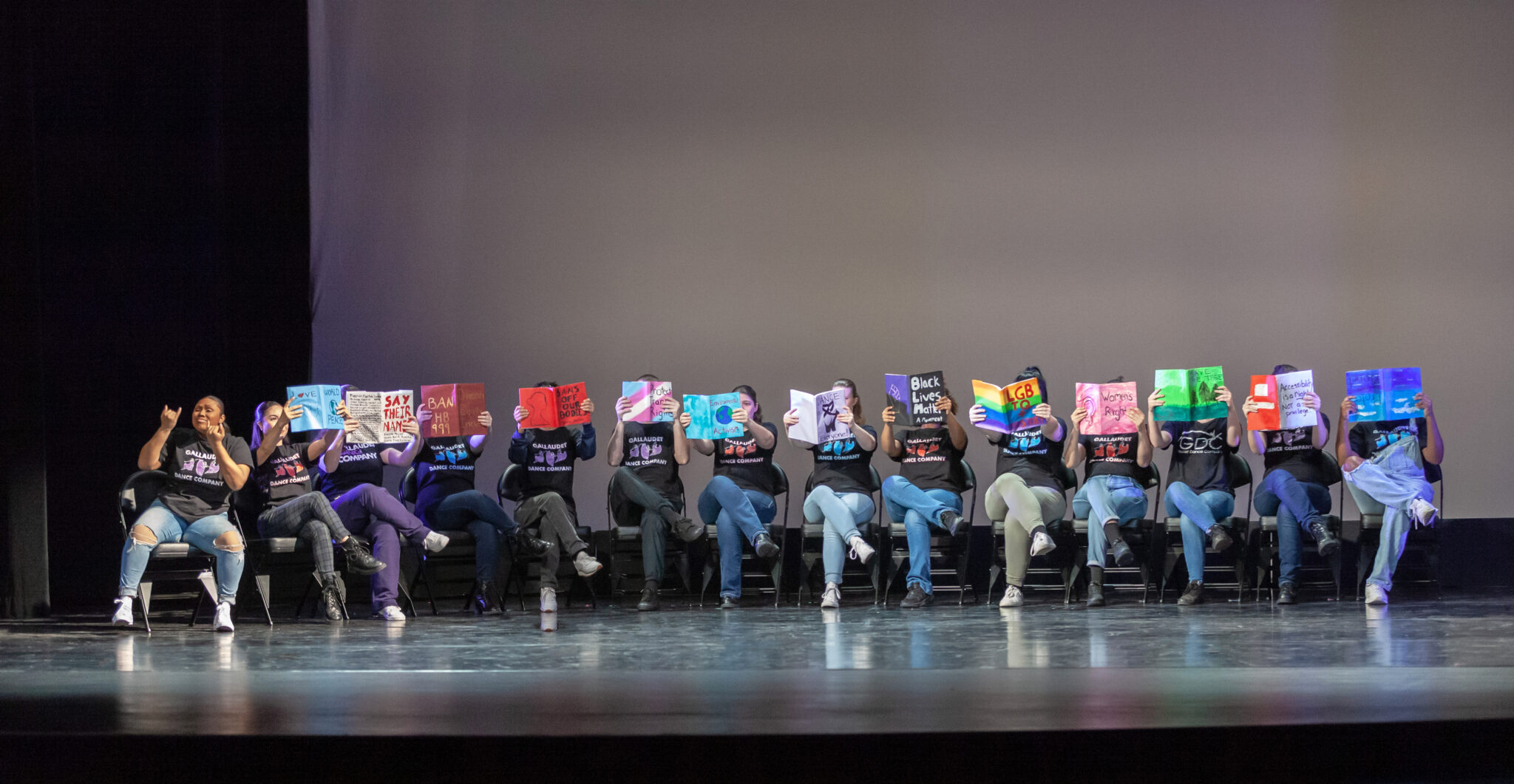 All dancers sitting in a line, one to the left signing and everyone else with a book on their face, all in GDC shirts and jeans
