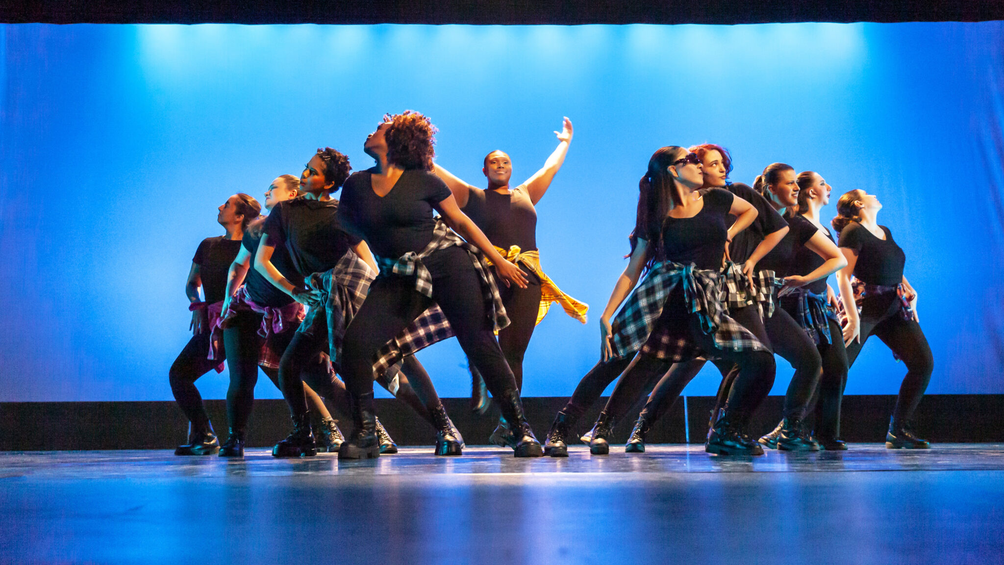 Blue background, dancers in all black with flannel on waits, o V shape split in the middle and dancer in the middle with hands up