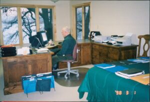 A man sitting at a desk in a home office, working on a computer. The office features large windows with a view of trees, wooden furniture, and various documents and folders on the desk. A green tablecloth is visible in the foreground, with some files stacked on it.