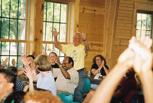 A lively group of people in a wooden room, with some individuals clapping and others raising their hands in enthusiasm. An older man in a yellow shirt stands at the back, cheering and celebrating.