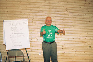 An older man wearing a green t-shirt is giving a thumbs-up gesture while standing next to a flip chart in a wooden-paneled room.