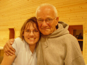A smiling woman with glasses and a light blue shirt embraces an older man wearing glasses and a gray hoodie, both standing in front of a wooden wall.