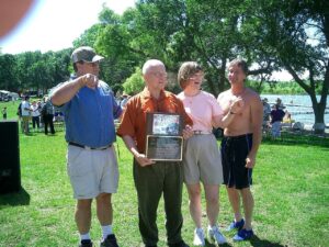 A group of four people standing outdoors, with a lake and trees in the background. One person, an elderly man in an orange shirt, holds a plaque. The others are casually dressed, with two men wearing shorts and a woman in a light-colored top. One man points while another smiles.