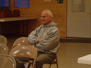 A person sitting in a chair in a room with wooden walls, wearing a gray hoodie and looking contemplative. Tables and chairs are arranged in the background, and there is a door visible on the right.