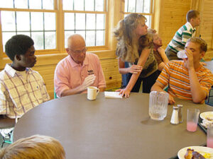 A group of people gathered around a table. Two young men are seated on the left, one with dark skin wearing a checkered shirt, and the other with light skin in a striped shirt. An older man in a pink shirt is sitting between them, holding a cup. A woman with long hair is leaning over the table, engaging with the group, while another young man in a striped shirt is sitting further back, drinking from a cup. The background features large windows with wooden walls.