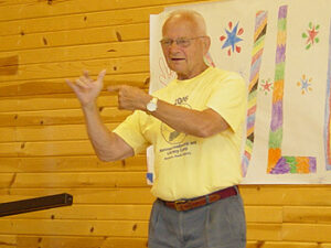 An elderly man wearing a yellow t-shirt is gesturing with his hands while standing in front of a wooden wall decorated with colorful drawings.