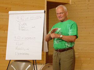 A smiling older man wearing a green t-shirt stands next to a flip chart. He is gesturing with his hands while speaking. The flip chart displays handwritten notes, including the titles "3 Rules of Education" and "3 Guidelines." The background features wooden walls, suggesting an indoor setting.