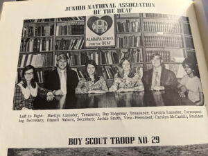 Black and white image of six individuals sitting at a table in front of a bookshelf. They are members of the Junior National Association of the Deaf, with a banner above them reading "ALABAMA SCHOOL FOR THE DEAF." The individuals are identified from left to right as Marilyn Lanaster, Bay Ridgway, Carolyn Lanaster, Dianne Nator, Jackie Smith, and Carolyn McCaul. The text at the bottom indicates "BOY SCOUT TROUP NO. 29."