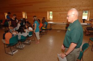 An older man in a green shirt stands to the side in a room with wooden walls, observing a group of people, mostly children and teens, seated at tables. Some individuals are interacting while others are engaged in activities. The atmosphere appears to be casual and communal.