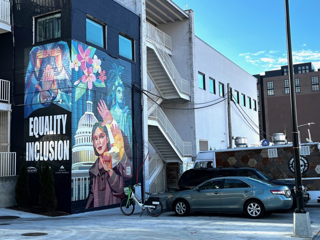 A colorful mural on the side of a building featuring the words "EQUALITY" and "INCLUSION." The mural includes a depiction of a woman with flowers, set against a backdrop that suggests a cityscape. In the foreground, a parked gray car and a bicycle are visible, along with architectural elements of surrounding buildings under a clear blue sky.