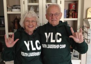 A smiling elderly couple wearing matching green hoodies with "YLC" and "YOUTH LEADERSHIP CAMP" printed on them, making hand gestures that resemble the sign for "I love you." They are standing in a cozy room with a shelf of decorative items in the background.