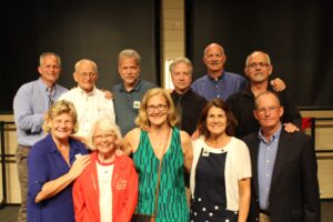 A group of eleven people poses for a photo on a stage, with a mix of men and women of varying ages. The individuals are standing and sitting in two rows, smiling at the camera. The background is dark, and the setting appears to be an indoor venue.