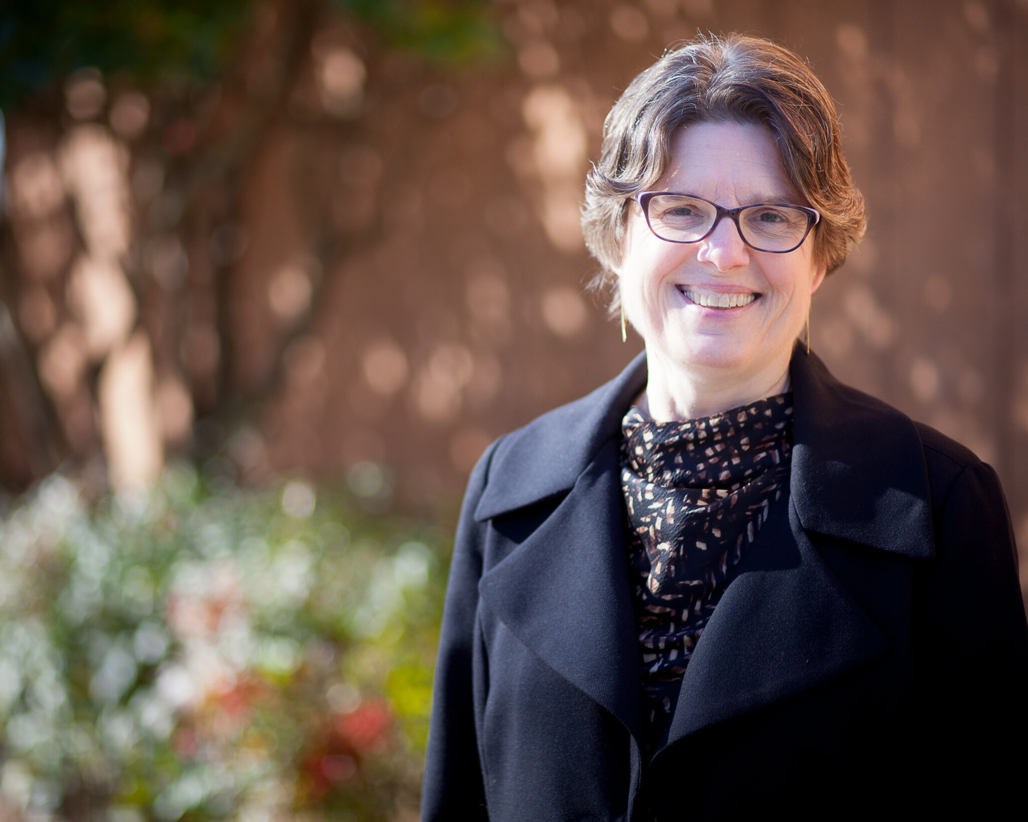 A woman with short, curly hair is standing outside, smiling. She is wearing glasses and a black coat over a patterned blouse. The background features soft, blurred greenery and warm sunlight.