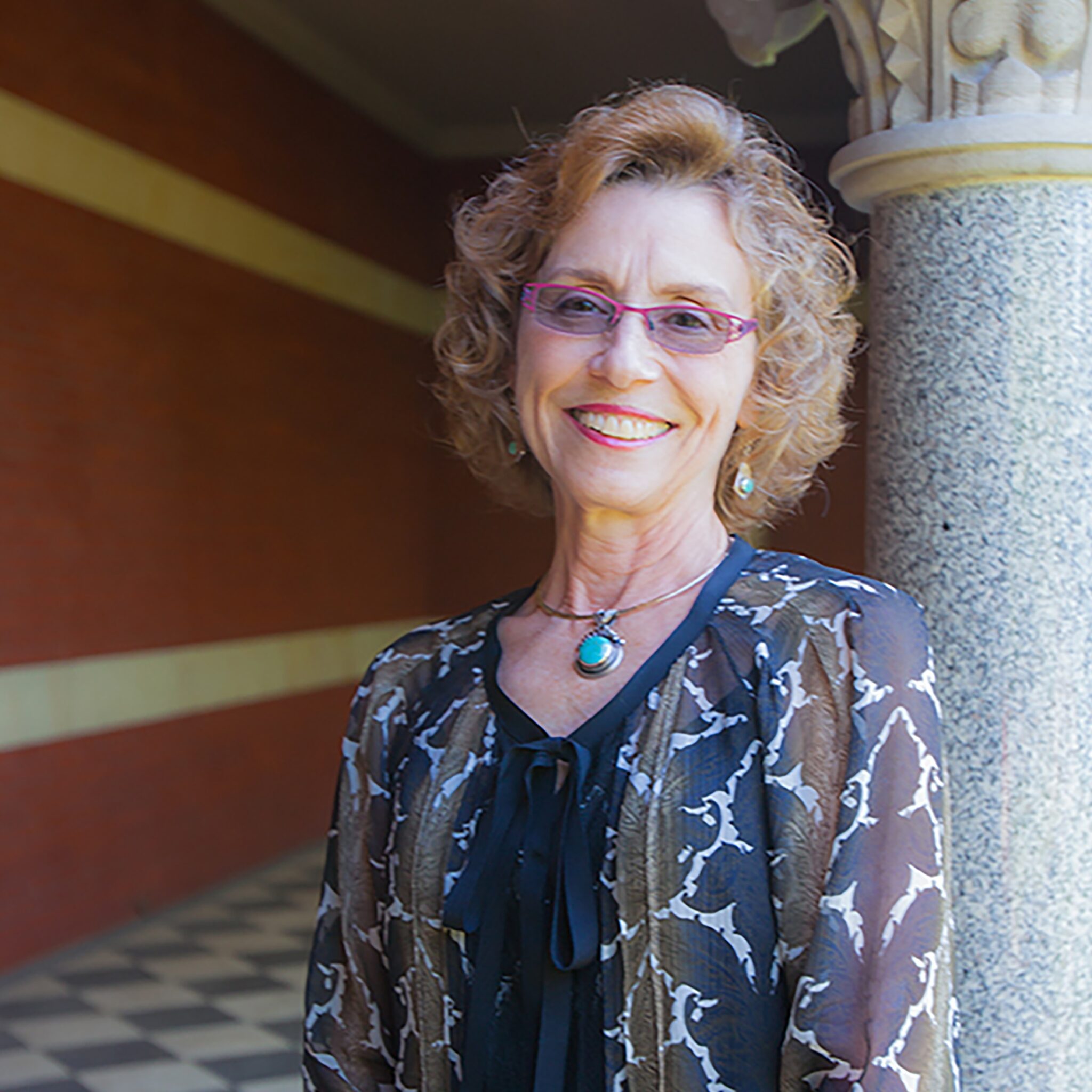 A smiling woman with curly hair, wearing glasses and a patterned blouse, stands near a stone pillar in a corridor with a checkered floor and red walls in the background.