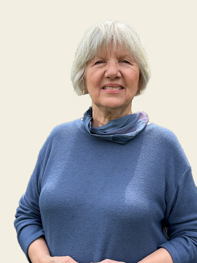 A smiling older woman with short, gray hair wearing a blue sweater and a light scarf, standing against a neutral background.