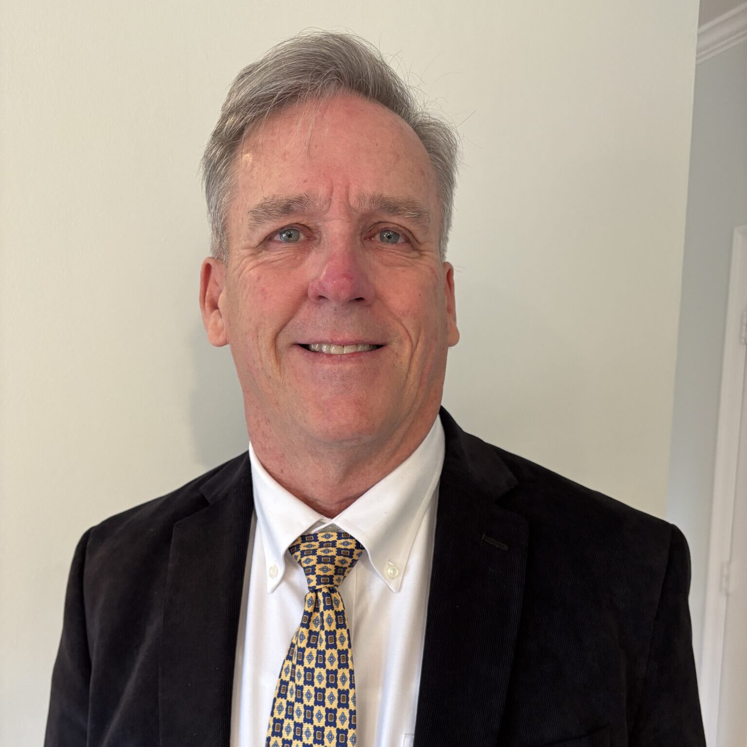 A man in a black blazer and white shirt, wearing a patterned tie, smiles at the camera. He has short, light-colored hair and is standing against a light-colored wall.