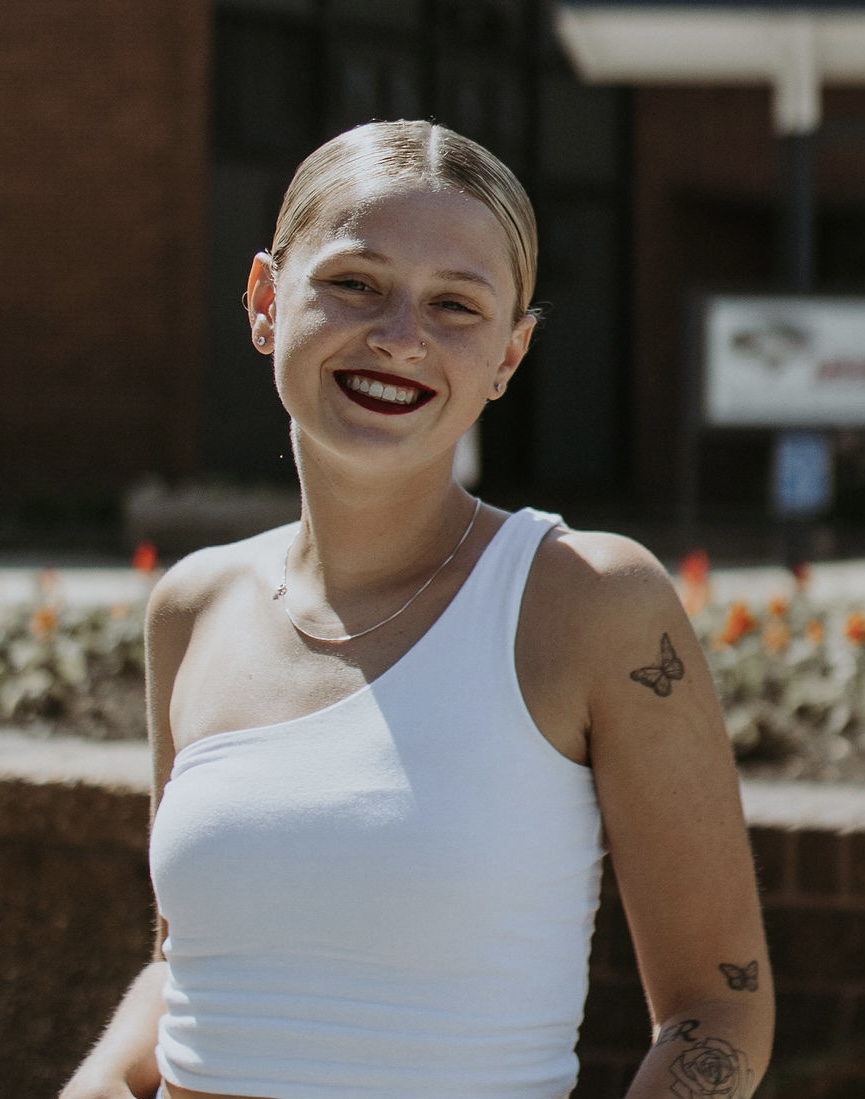 A young person with short, light-colored hair smiles brightly, wearing a sleeveless white top. They have a butterfly tattoo on their arm and are standing in front of a brick wall with flowers in the background.