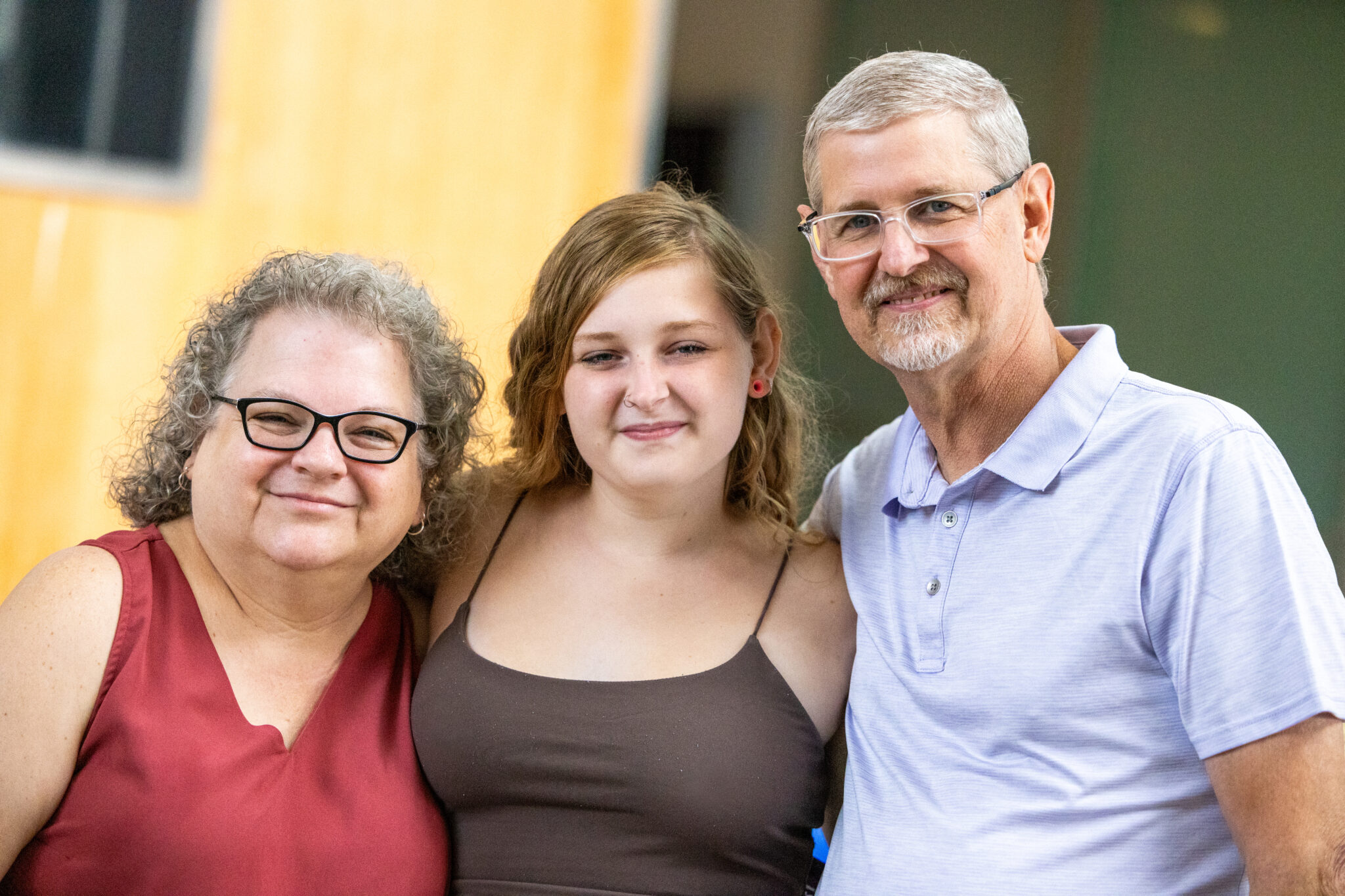 A smiling woman with curly gray hair and glasses stands on the left, a young woman with wavy brown hair in the middle, and a man with short gray hair and glasses on the right. They are standing close together outdoors, with a blurred background featuring warm colors.