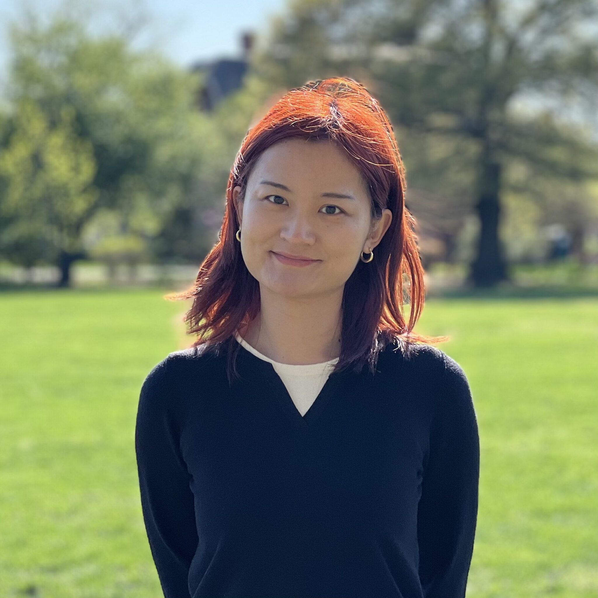 A young woman with shoulder-length reddish hair stands smiling in a park. She is wearing a dark sweater over a white shirt, with green grass and trees in the background, under a clear blue sky.