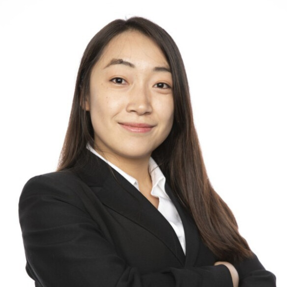 A professional woman with long dark hair, wearing a black suit and white shirt, stands confidently with her arms crossed and a subtle smile against a plain white background.