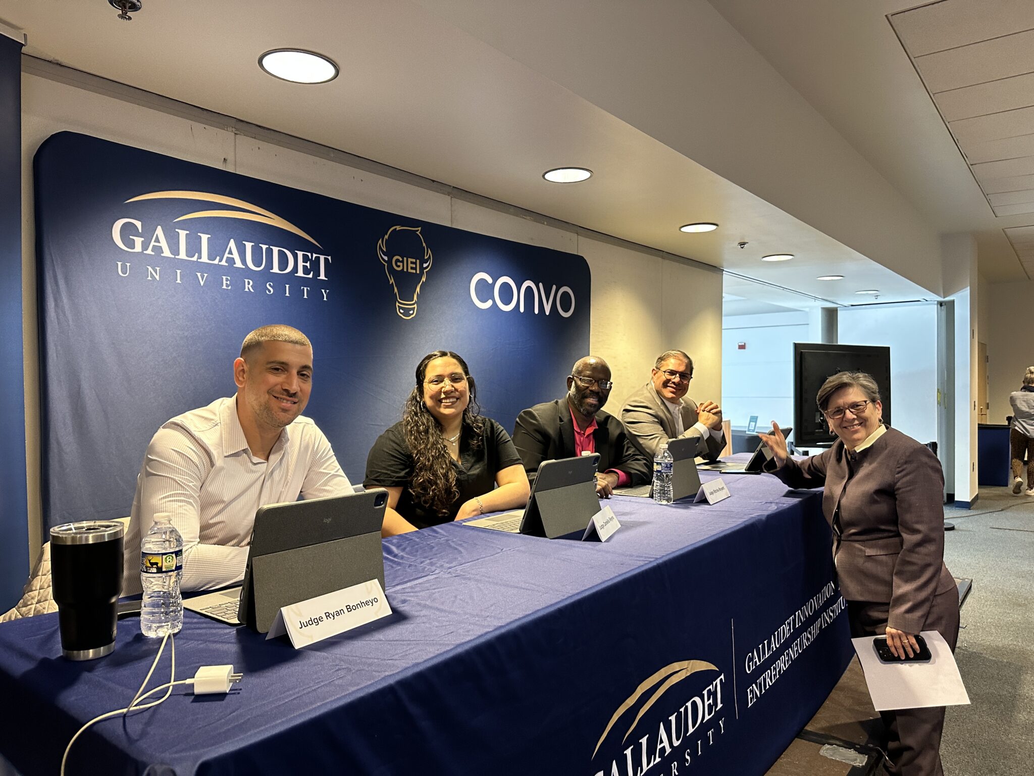Four people sit behind a long blue table. Each has a computer and name placard in front of them. On the other side of the table is a woman standing up and gesturing toward the camera.