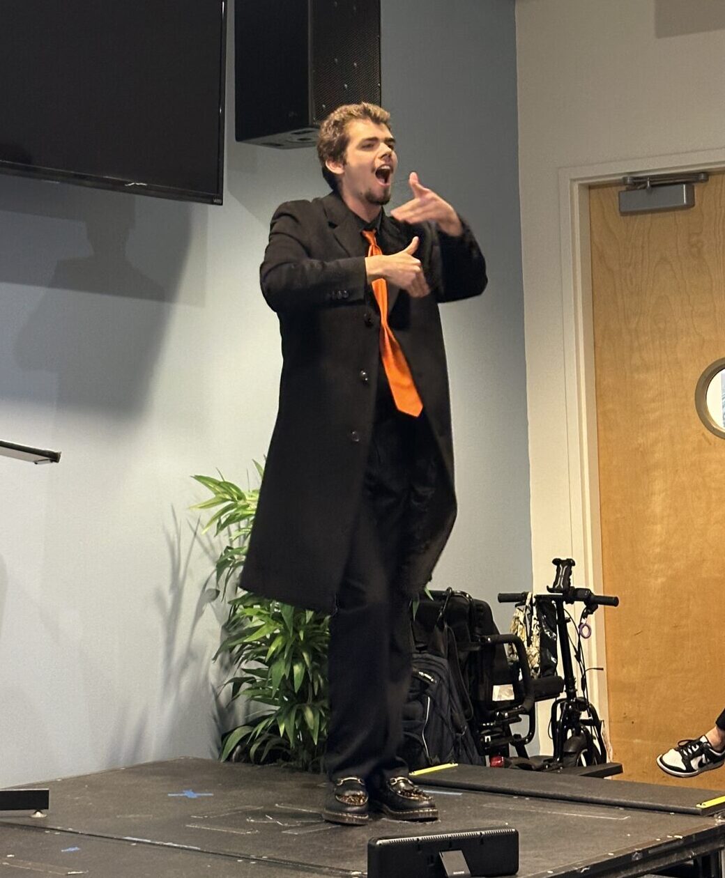 Man in black with a long orange tie signs passionately while on a stage.