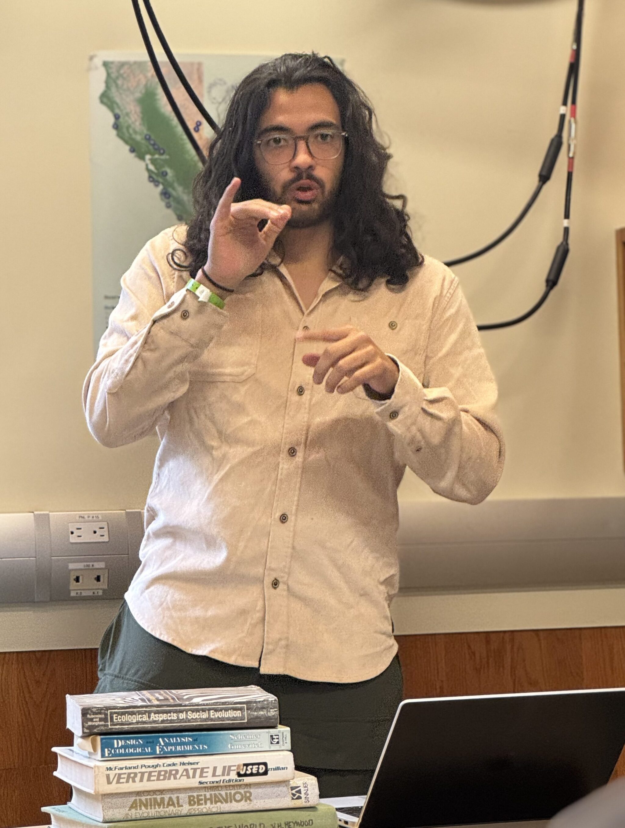Man with long hair signs while standing in a room with white walls. Behind him is a map of California. On a table in front of him is a stack of books.