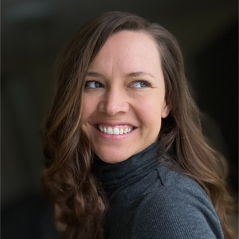 A woman with long, wavy brown hair is smiling while looking over her shoulder. She is wearing a dark turtleneck sweater and has a soft, warm expression on her face. The background is softly blurred, creating a focus on her features.