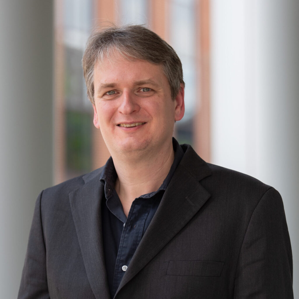 A photo of Dr. Christian Vogler smiling at the camera. He has short, light brown hair. He is wearing a dark blazer over a dark button-up shirt. The background is an out-of-focus building element that could be an entryway.